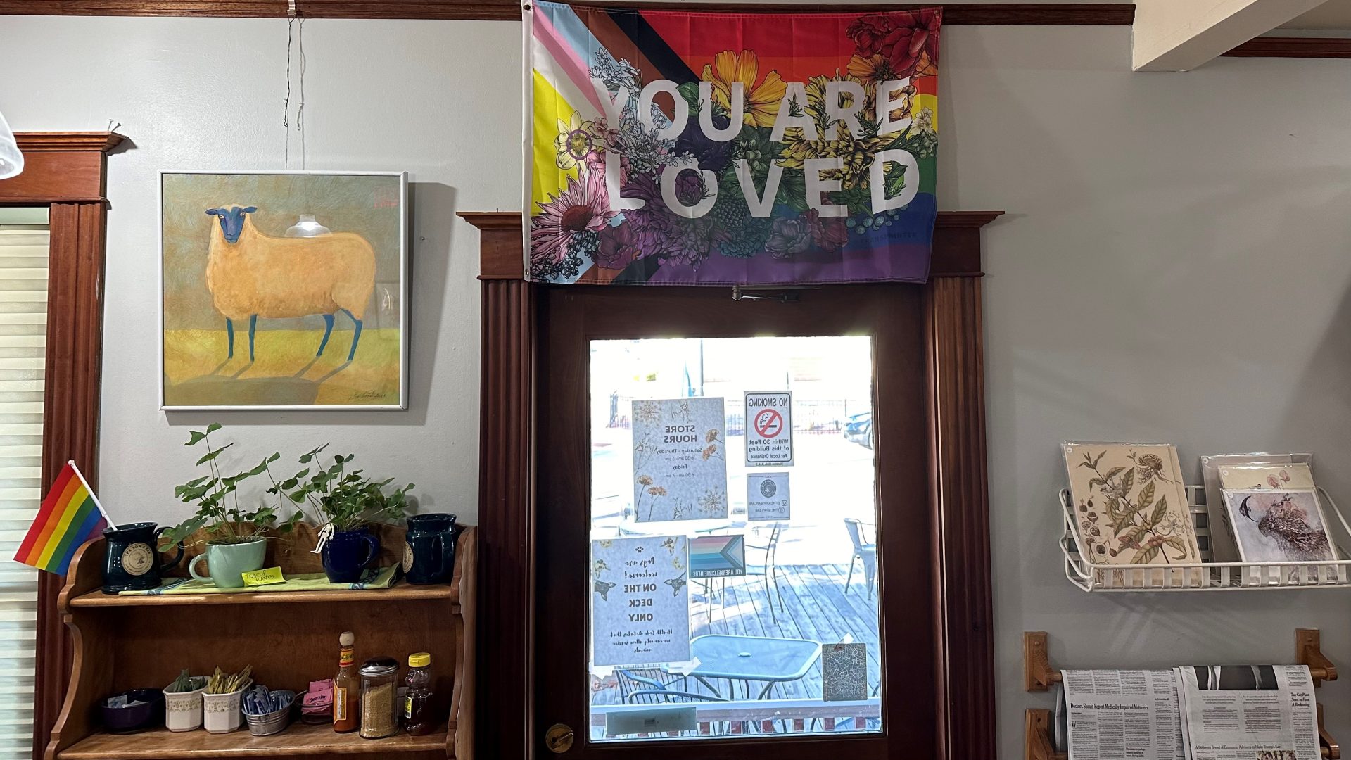 The flag that hangs over the door at the Sow's Ear as a message to our community: it is the progress pride rainbow flag overlaid with flowers and the words YOU ARE LOVED.