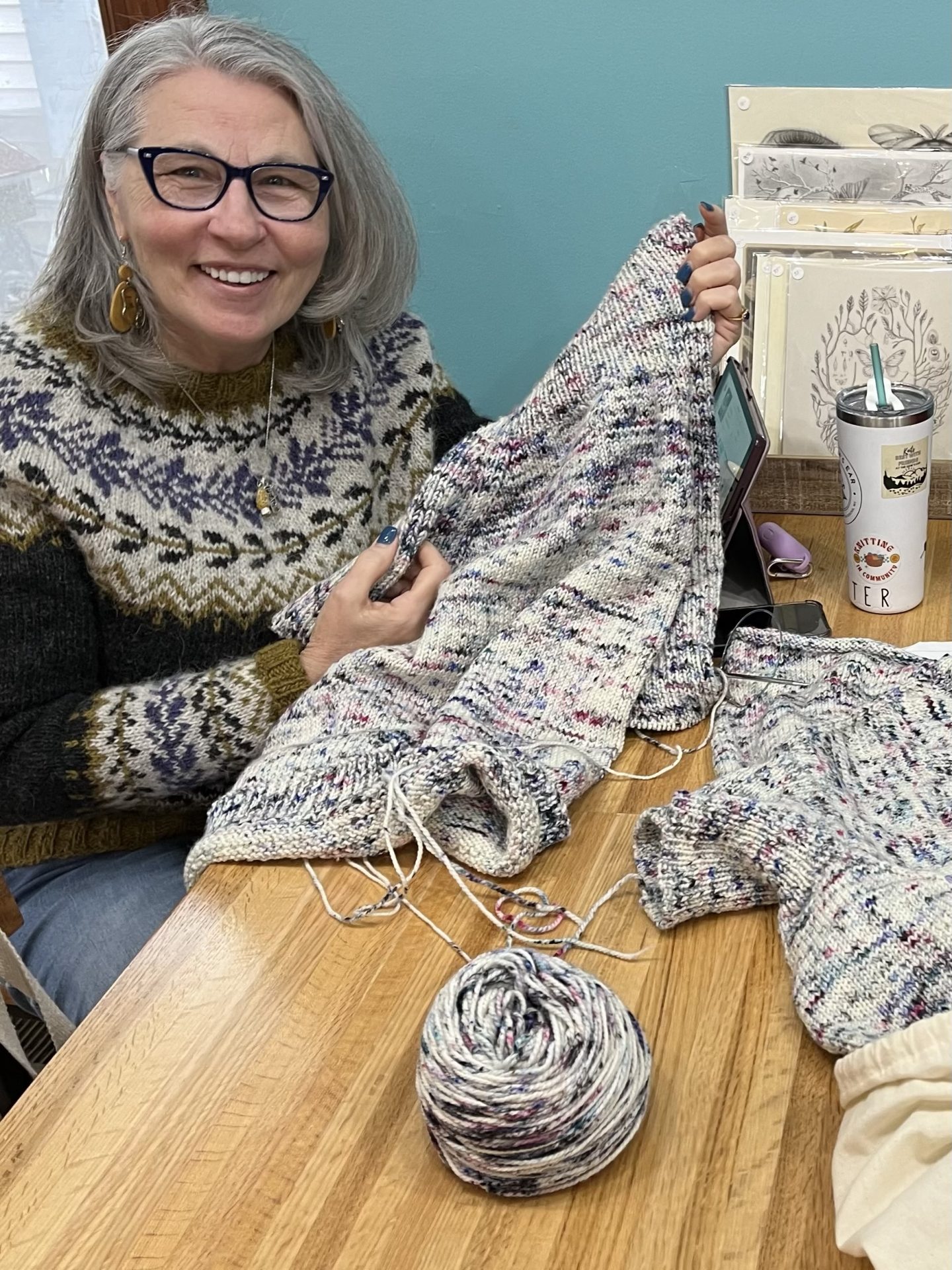 Jean Marie smiling and holding up a lovely almost-completed knit sweater while wearing another handknit sweater decorated with vine and flower motifs.