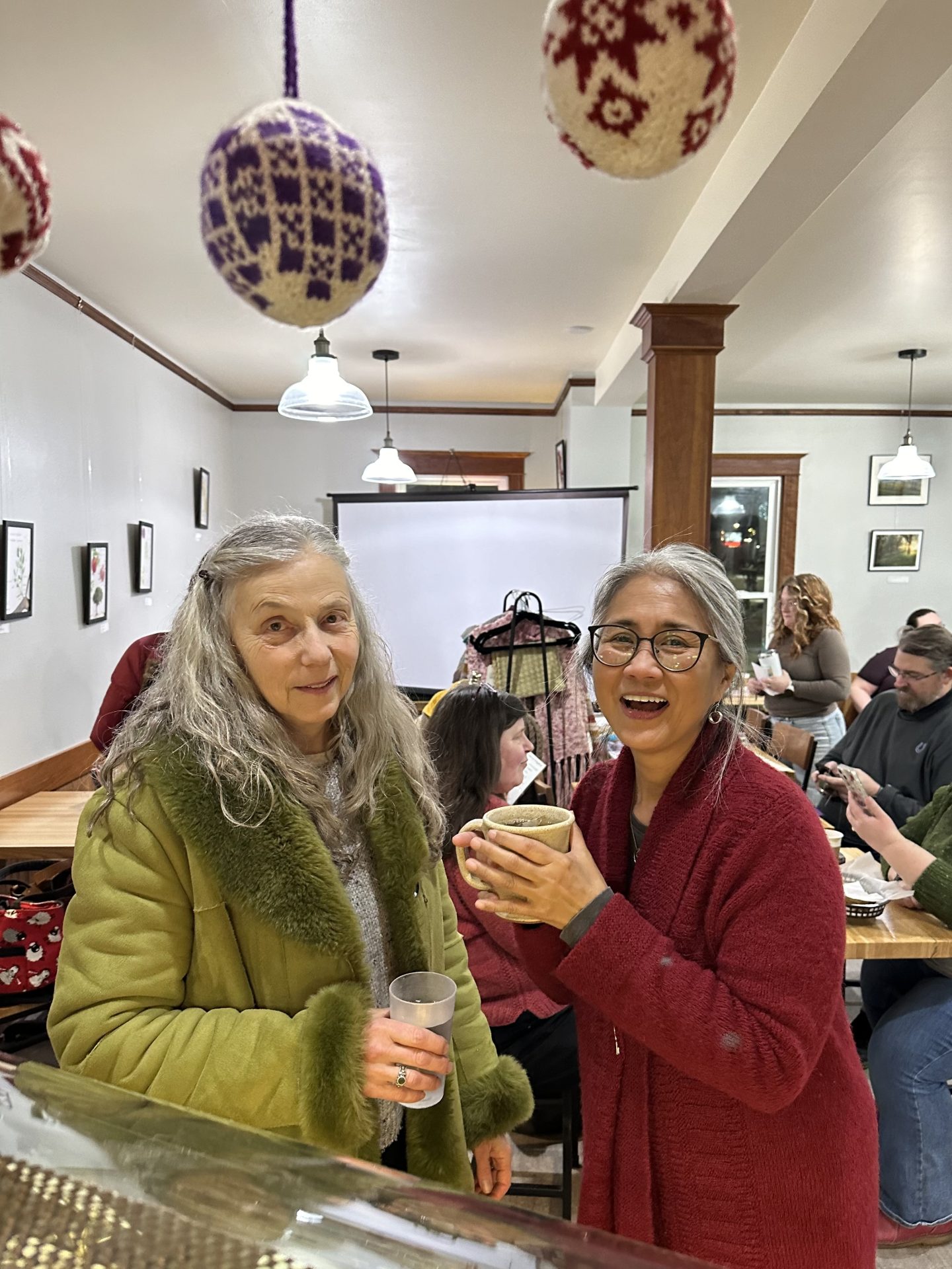 Melissa Brown in a long green coat and Joan Looi in a red handknit sweater, smiling in the Sow's Ear cafe during an event.